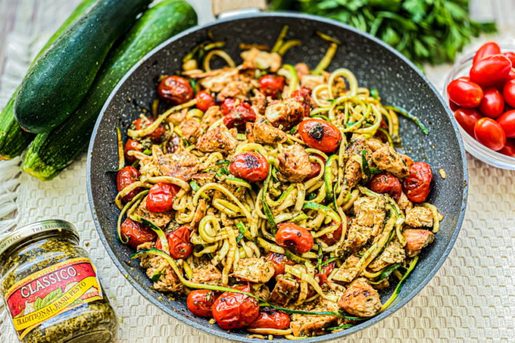 Our One Pan Chicken Zoodles in a skillet next to a jar of pesto, zucchini, and cherry tomatoes.