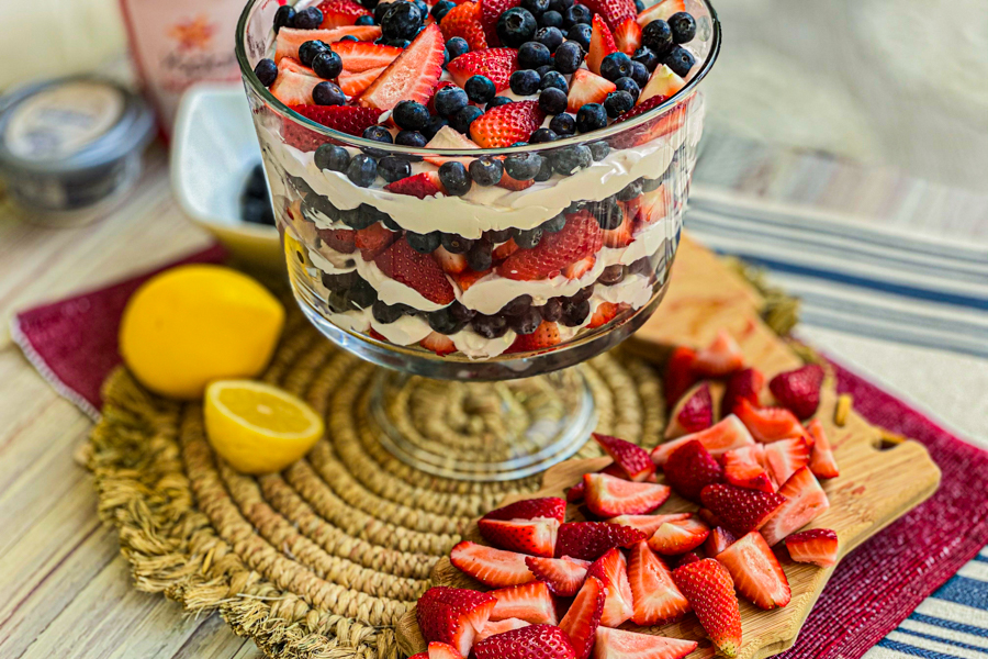 Our Easy Berry Trifle all finished next to a cutting board of cut strawberries, a sliced lemon, and a bowl of blueberries. 