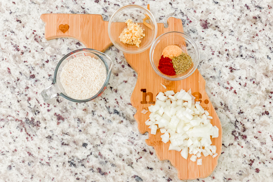 A florida-shaped cutting board with diced onions, diced garlic, and a small bowl of seasoning on top. 