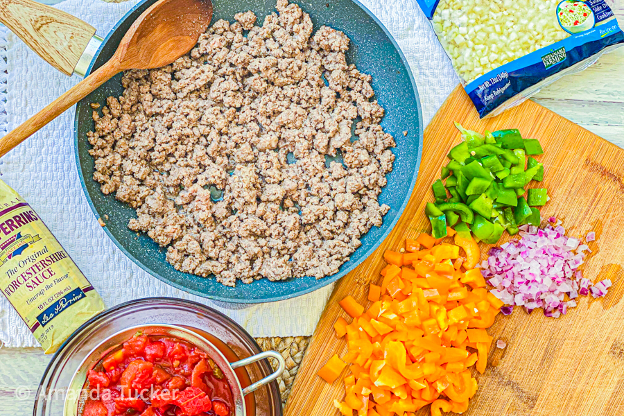 A pan of browned ground beef next to a cutting board with chopped onions and bell peppers on it. 