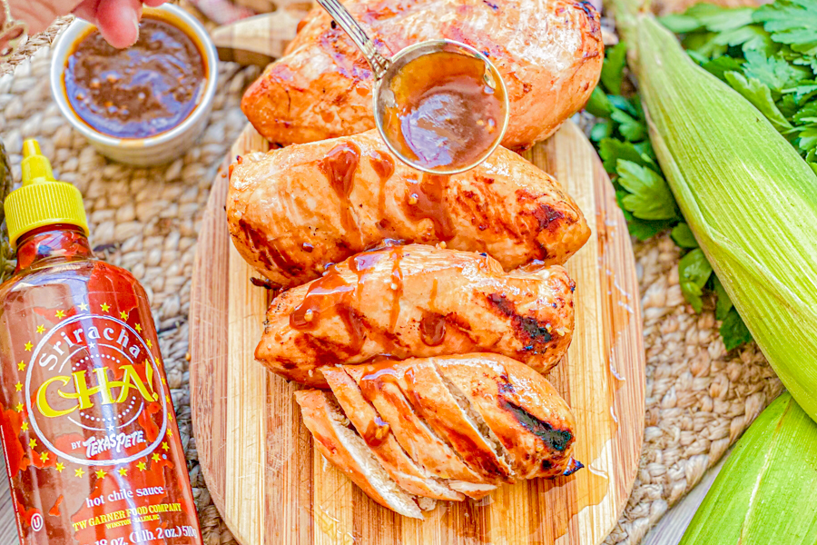 Grilled chicken on a cutting board next to a bottle of Sriracha sauce. 