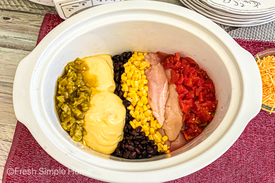 A white crockpot bowl with the Rotel, black beans, corn, chicken, green chiles, and cream of chicken soup in it. 